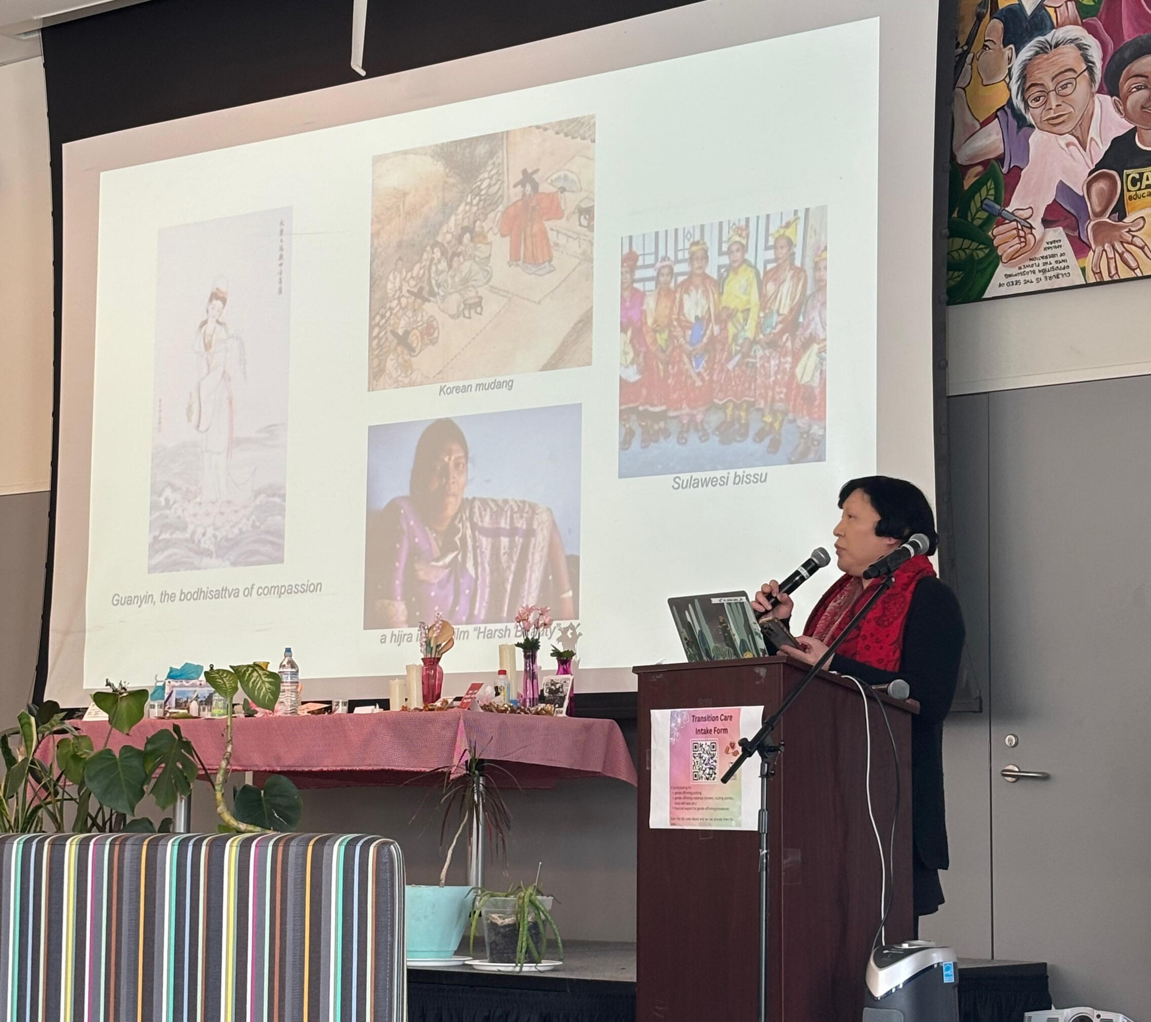 Transgender activist Pauline Park delivering keynote address at QACON 2025 An Asian woman with short black hair stands at a podium speaking into a microphone. Behind her is a slide show showing pictures of guanyin, Korean mudang, sulawesi bissu, and a hijra
