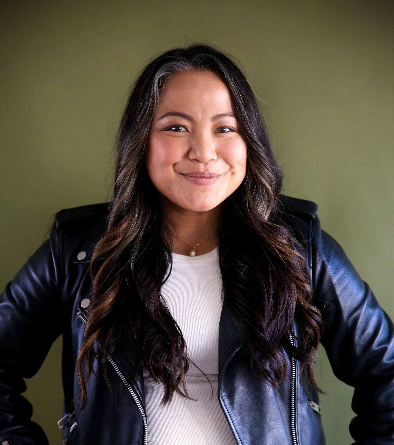 Asian woman with long wavy dark brown hair smiling and wearing a dark leather jacket and white shirt