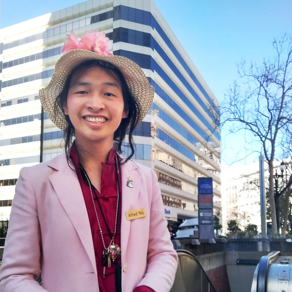 Person with long black hair and medium light skin tone wears a red colored shirt, pink jacket, and wide-brimmed hat with pink flowers on it, standing in front of BART subway entrance