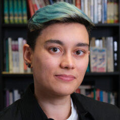 Person with medium skin and short dark hair died teal blue looks straight at camera with bookshelves in the background