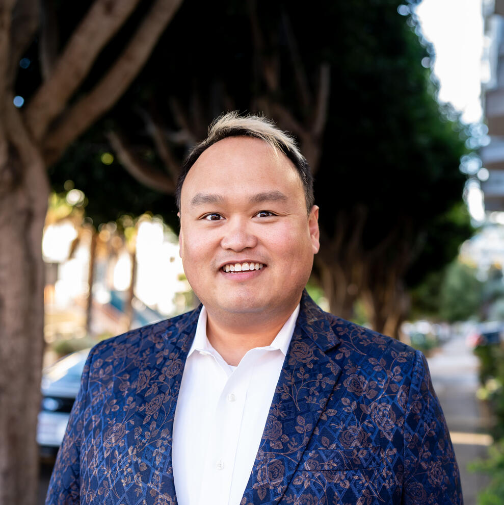 Michael Nguyen Asian person with short dark hair that is light blonde at the ends smiles in front of trees on a city street, wearing a white colored shirt and blue rose patterned jacket
