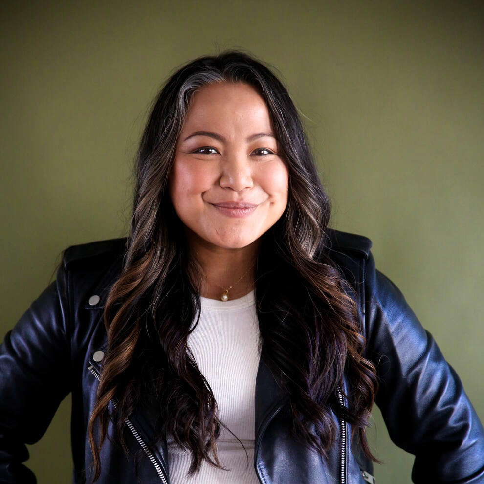 Asian woman with long wavy dark brown hair smiling and wearing a dark leather jacket and white shirt