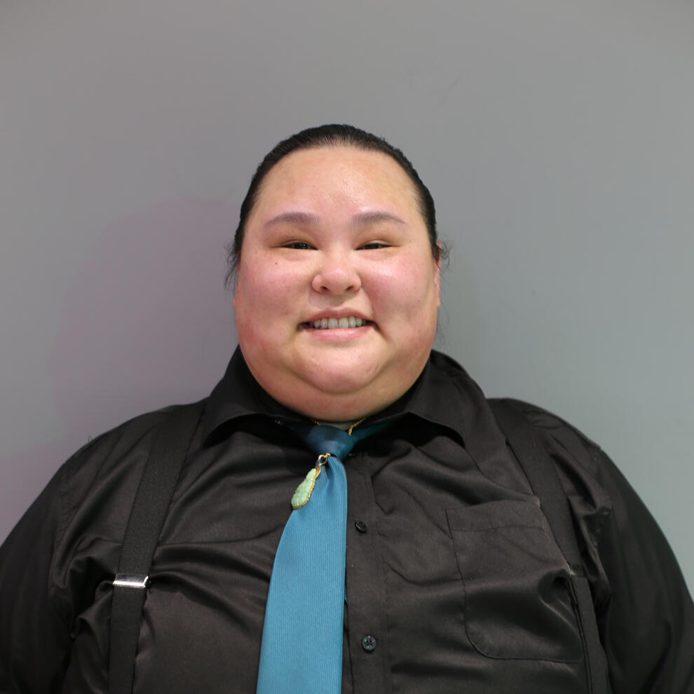 Person with medium skin and dark hair that is tied back smiles at camera wearing a dark collared shirt, teal tie, and black suspenders.